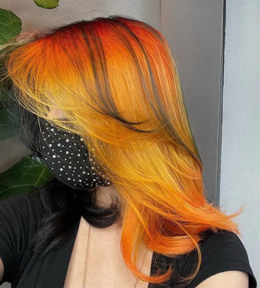 A person with a layered balayage hairstyle featuring bright orange and yellow color with black undersides, wearing a black rhinestone face mask. - Ka'ton Salon, Fort Lauderdale FL