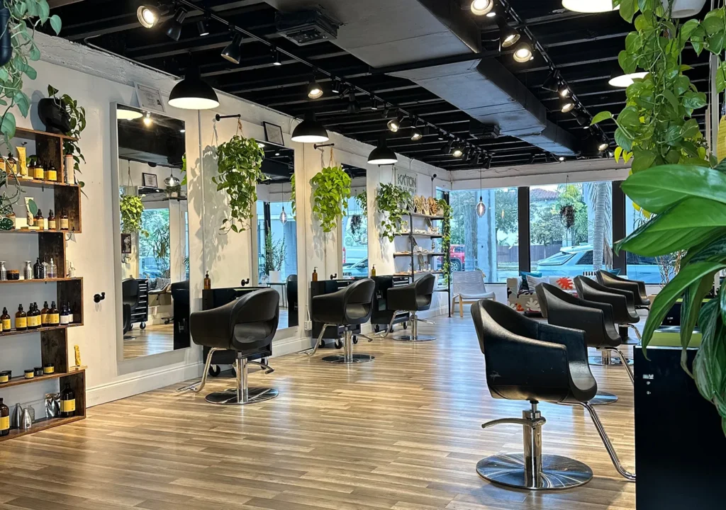 Interior of a hair salon for a haircut, featuring black styling chairs, mirrors, shelves of hair products, and hanging potted plants. - Ka'ton Salon, Fort Lauderdale FL