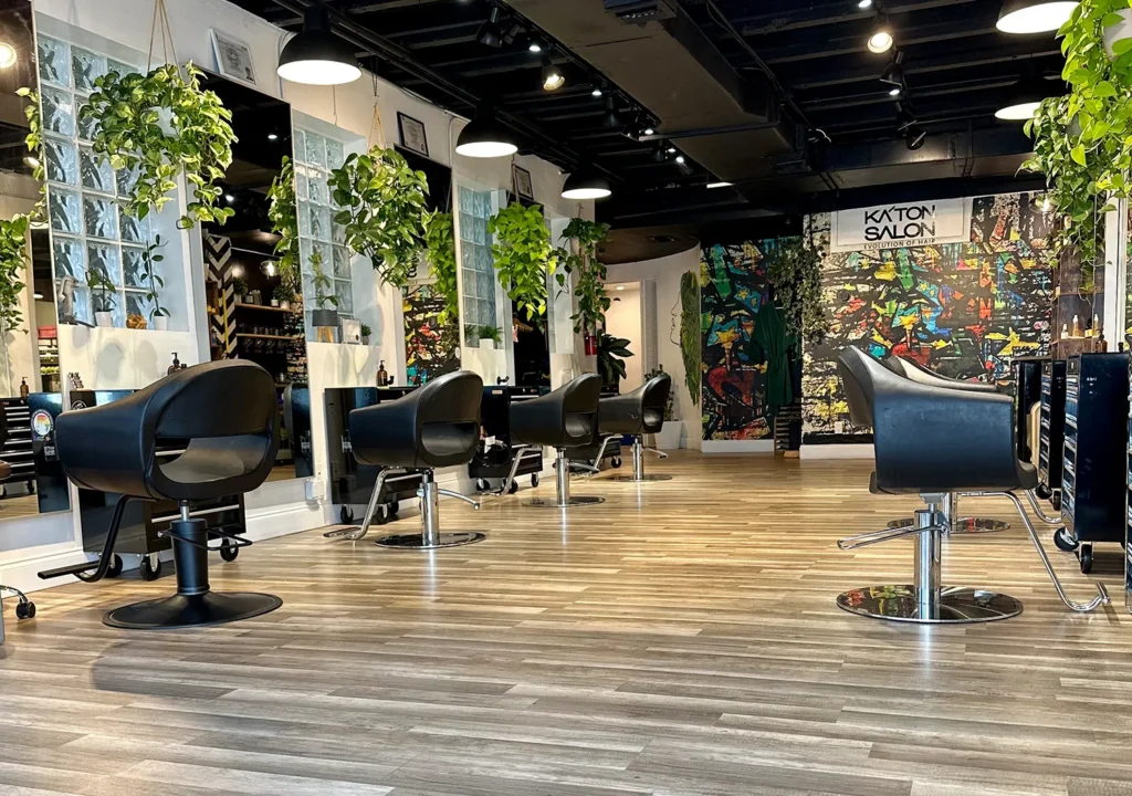 Interior of a hair salon with rows of black styling chairs for a haircut, hanging plants, and a colorful abstract mural on the back wall. - Ka'ton Salon, Fort Lauderdale FL