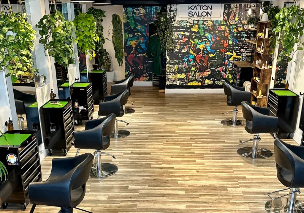 Interior of a hair salon with black styling chairs for a haircut or blowout, rolling carts, hanging plants, and a colorful graffiti-style mural on the back wall featuring the Katon Salon logo. - Ka'ton Salon, Fort Lauderdale FL