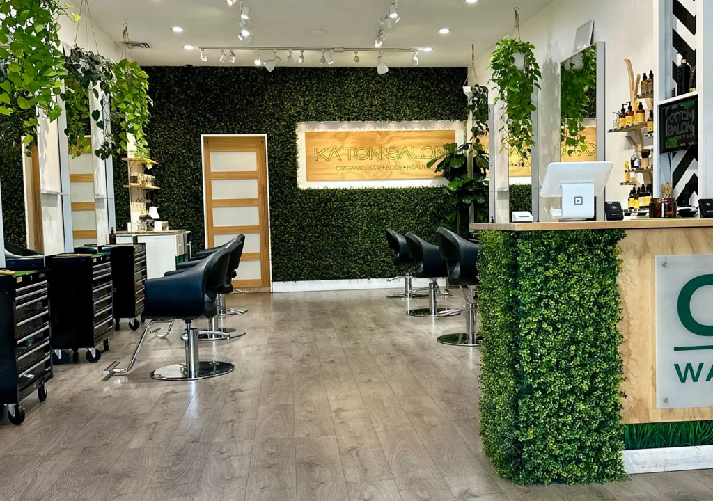 Interior of Katon Salon featuring rows of hair stylist chairs, hanging plants, and a greenery-covered reception desk with a sign reading “Katon Salon Organic Hair Body Health.” - Ka'ton Salon, Fort Lauderdale FL