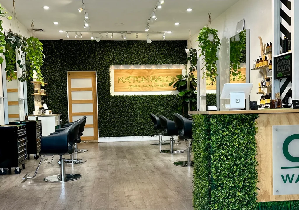 Interior of Katon Salon with rows of black styling chairs for a blowout, a green artificial grass wall with a wood-mounted sign, and a reception desk covered in greenery. - Ka'ton Salon, Fort Lauderdale FL