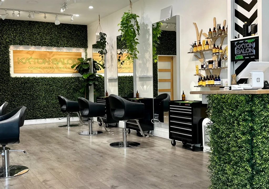 Interior of a hair salon with black styling chairs, a reception desk, and a wall sign for Katon Salon, which specializes in haircut and balayage services for organic hair, body, and health. - Ka'ton Salon, Fort Lauderdale FL