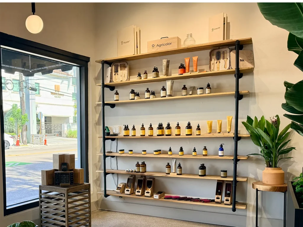 Shelves mounted on black pipes display hair care products for a blowout, with a small wooden crate of merchandise near the window. - Ka'ton Salon, Fort Lauderdale FL