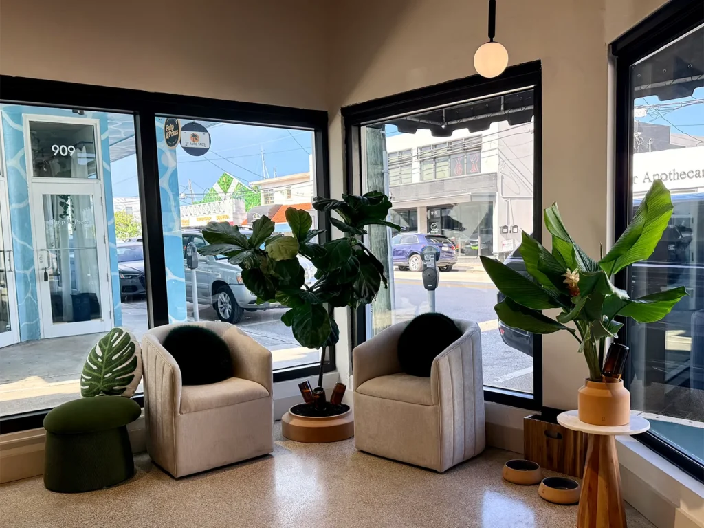 A seating area for a hair salon with two beige armchairs, a large fiddle leaf fig tree, and a smaller potted plant in front of floor-to-ceiling windows. - Ka'ton Salon, Fort Lauderdale FL