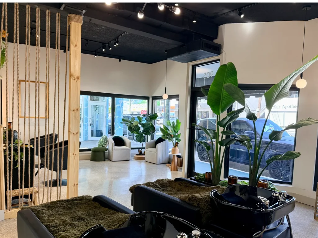 Interior of a hair salon with two black shampoo bowls for a blowout in the foreground and a seating area with chairs and potted plants by the windows in the background. - Ka'ton Salon, Fort Lauderdale FL